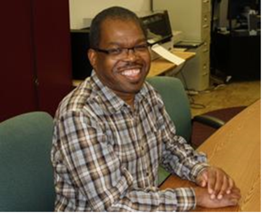 Image of Herschel Jackson, a Black man wearing glasses and a plaid shirt sitting in a desk chair and smiling for the camera.