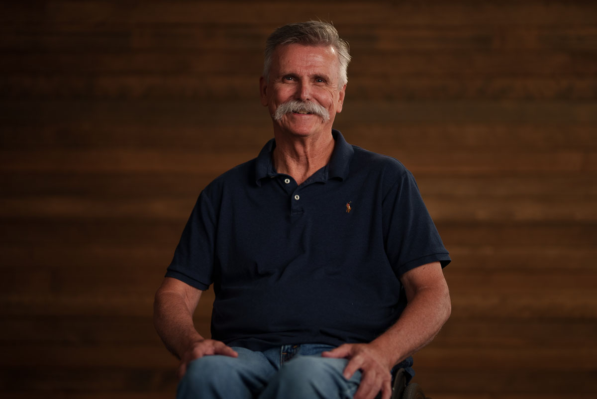 Photo of Scott Birkenbuel, a white man with salt and pepper mustache and hair. He is sitting with hands by his side in a relaxed position wearing a black short-sleeve collared shirt and jeans.
