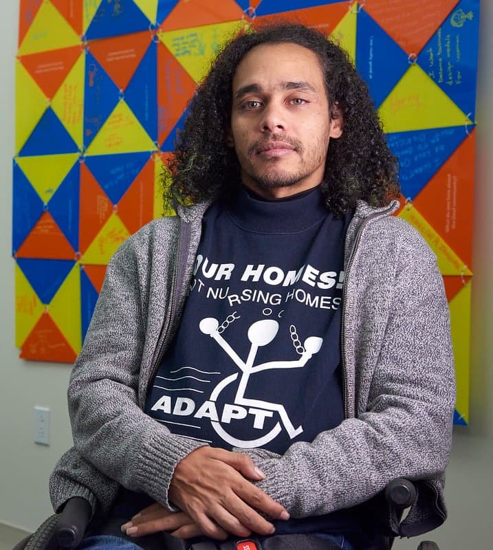 Full front photo of Germán Parodi. A man sitting in his wheelchair with a slight grin and hands crossed on his lap. He has brown skin, and shoulder length curly dark brown hair. He is wearing a black long sleeve shirt that says “Our Homes Not Nursing Homes ADAPT ” on it and a gray zip-up sweater on top. Behind him appears a painting with multi-colored triangles.   