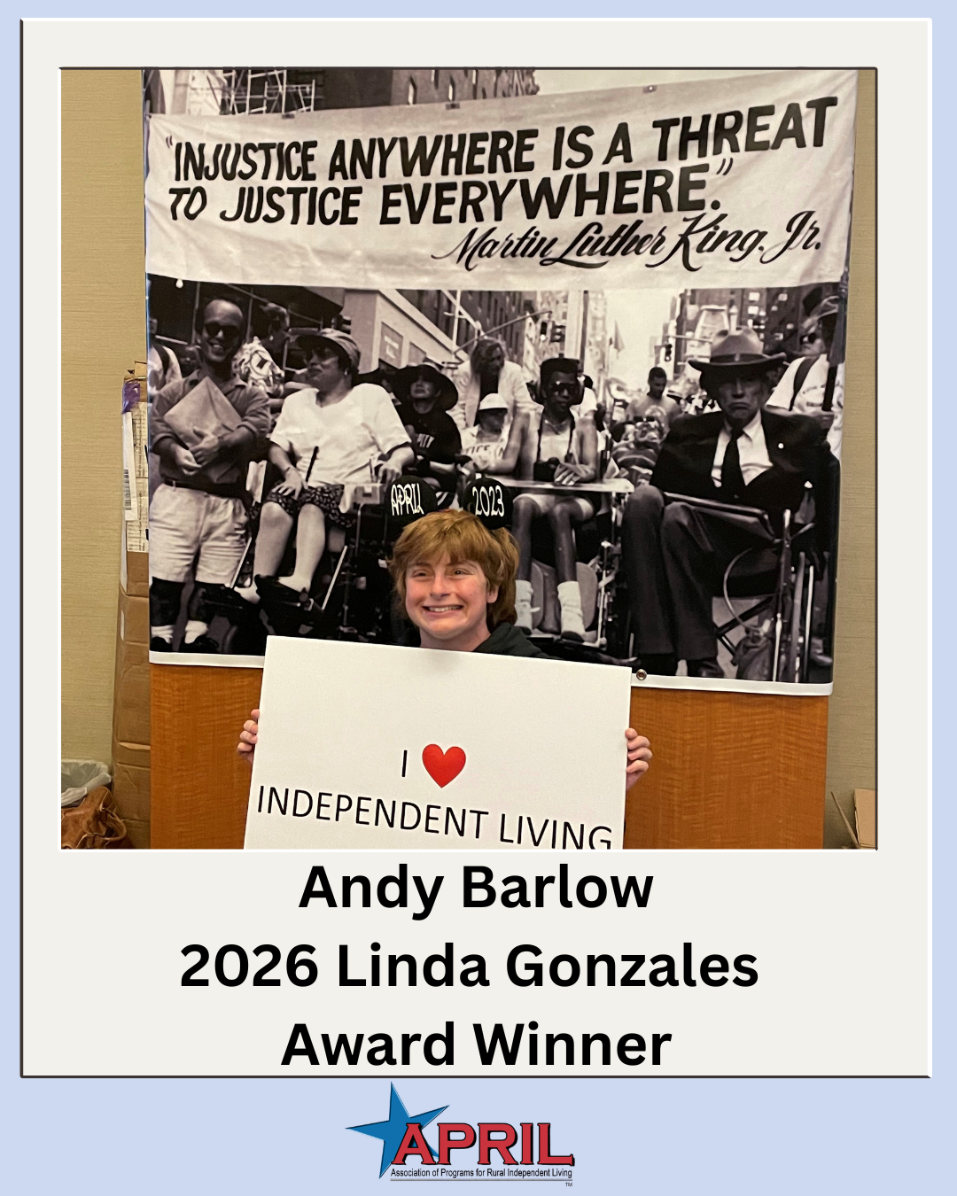 Photo of Andy Barlow holding a "I HEART independent living" sitting in front of a backdrop of disability rights activists protesting with a large banner sign that says "INJUSTICE ANYWHERE IS A THREAT TO JUSTICE EVERYWHERE. - MARTIN LUTHER KING, JR".