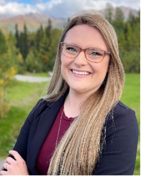 Photo of Brigit Barstad, a young white woman with long blonde hair and dark rimmed glasses posing for a photo outside.