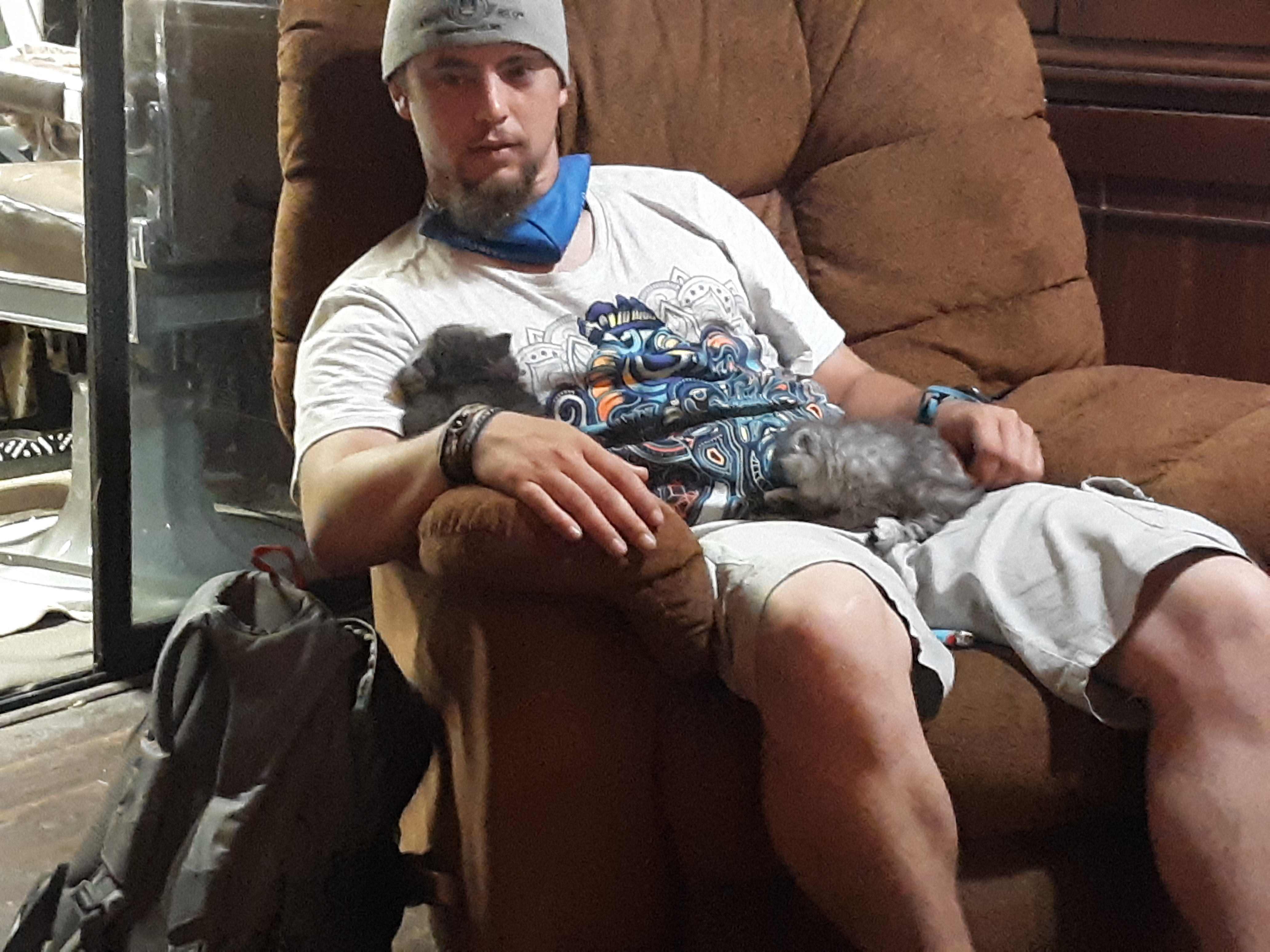Photo of Evan Shockley. Evan is sitting on an oversized brown couch with two persian cats on his lap. He is wearing shorts and a t-shirt and beanie. Evan describes this photo as "chillin' with the persians".