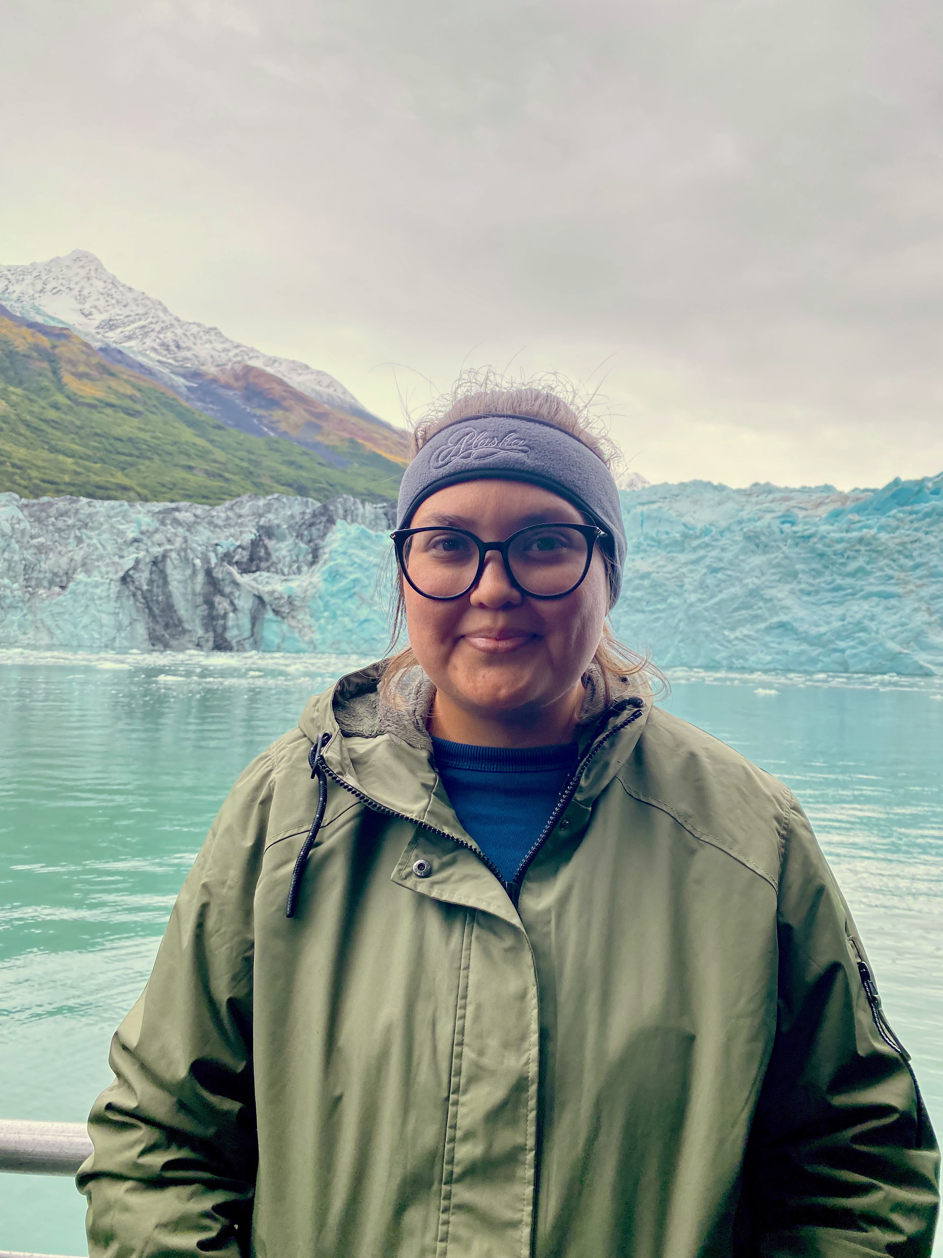 Photo of Anya Carillo, an Indigenous woman. She is standing in front of a body of water and mountains, wearing a green coat, glasses, and headband.