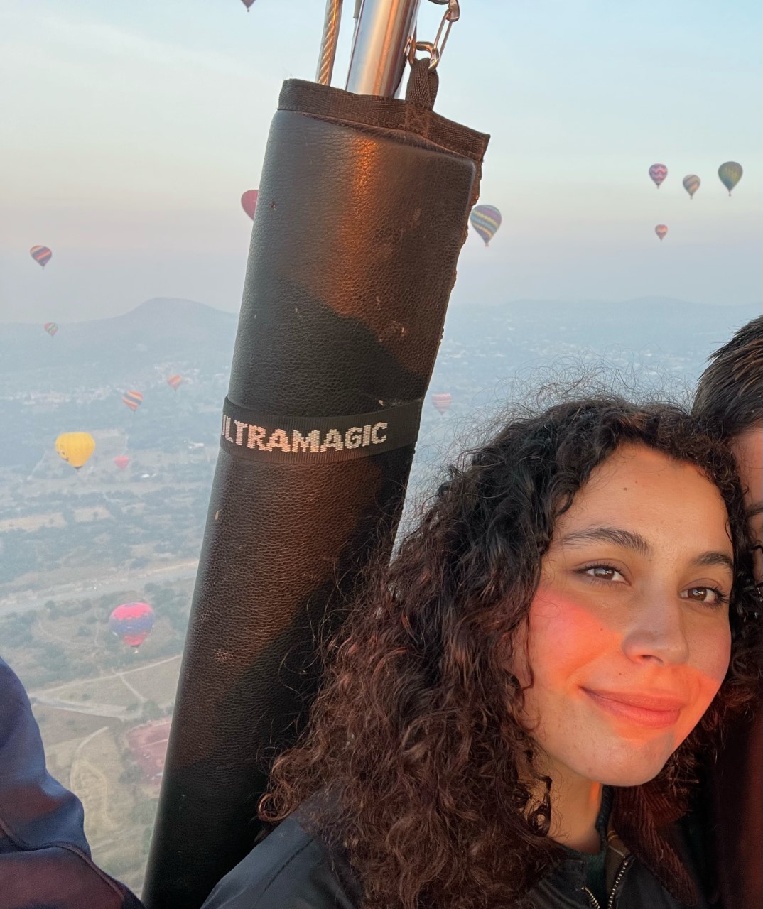 Photo of Jazmin Barajas in a hot air balloon. She has long curly dark hair and is smiling at the camera with hot air balloons in the distance.