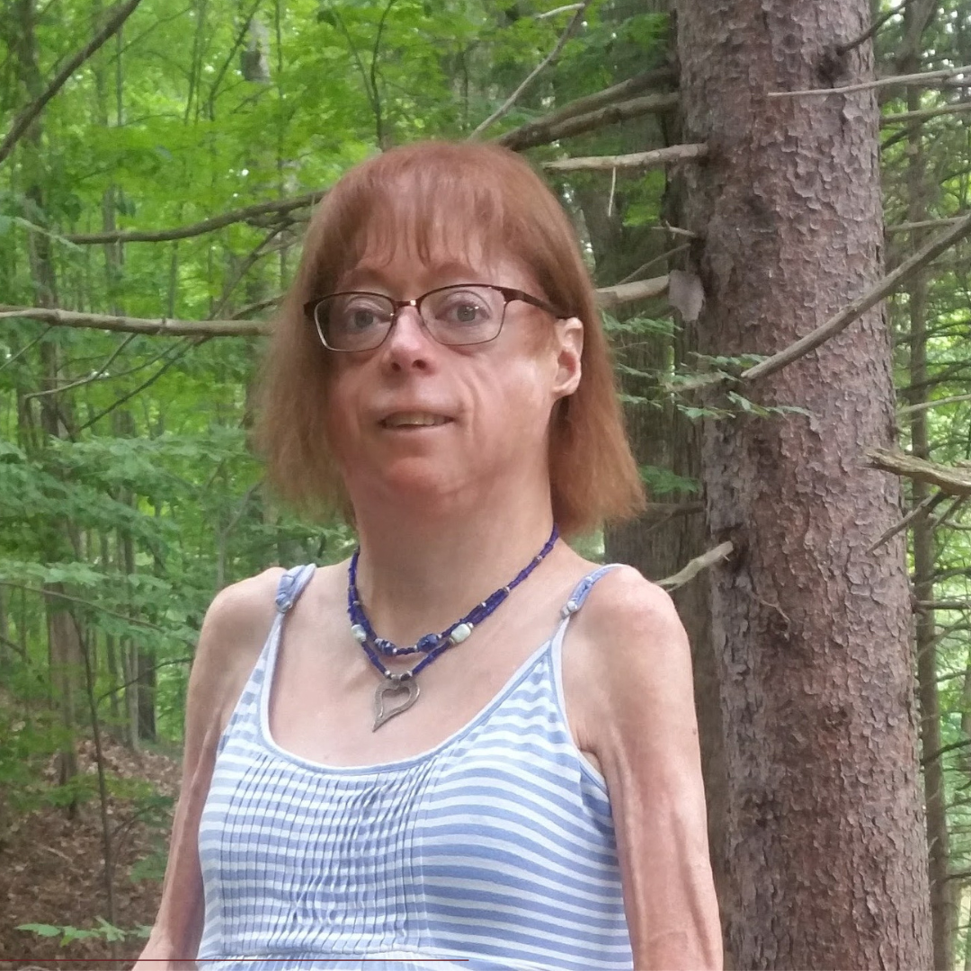 Image of Joan Labelle, a white woman with red shoulder length hair and a bangs. She is wearing glasses and a striped tank top standing outside with trees behind her.