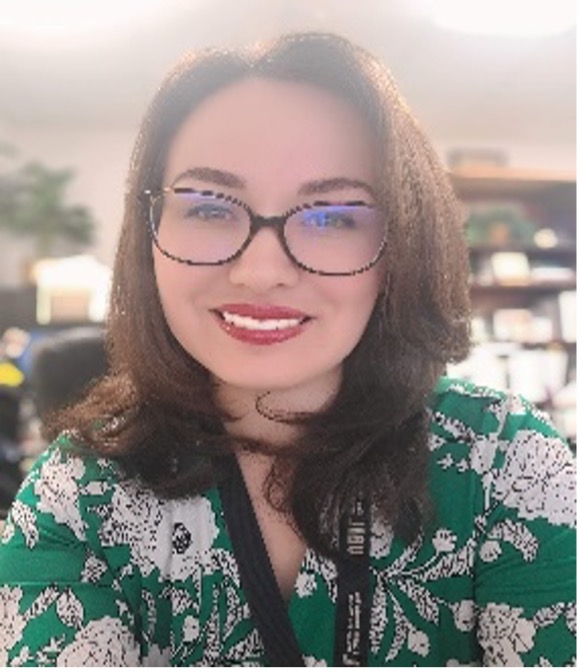 I have fair skin and I am a Hispanic Woman in her mid-30s. Wearing large black rim glasses and a green blouse with white flowers printed on it. My hair is brown, and it is straight and shoulder length. I have green eyes and I am wearing red lipstick. I am sitting in my office with a bookcase behind me. My preferred pronouns are She/Her/Hers.