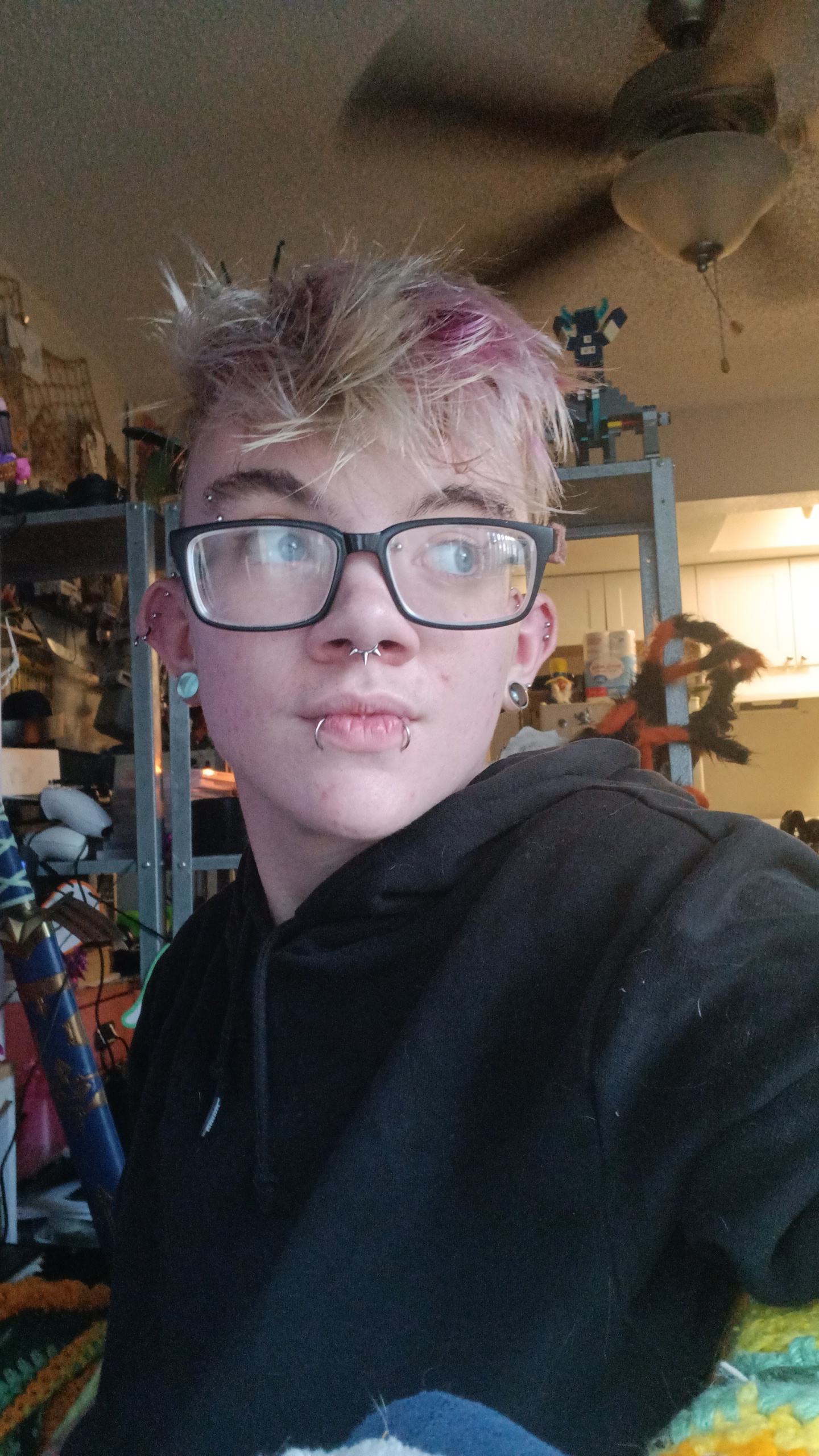 Photo of Maxx Gastelum, a young adult with dissheveled blonde hair with a streak of faded pink in it. amaxx is wewaring glasses, has multiple facial piercings, and is wearing a black top.