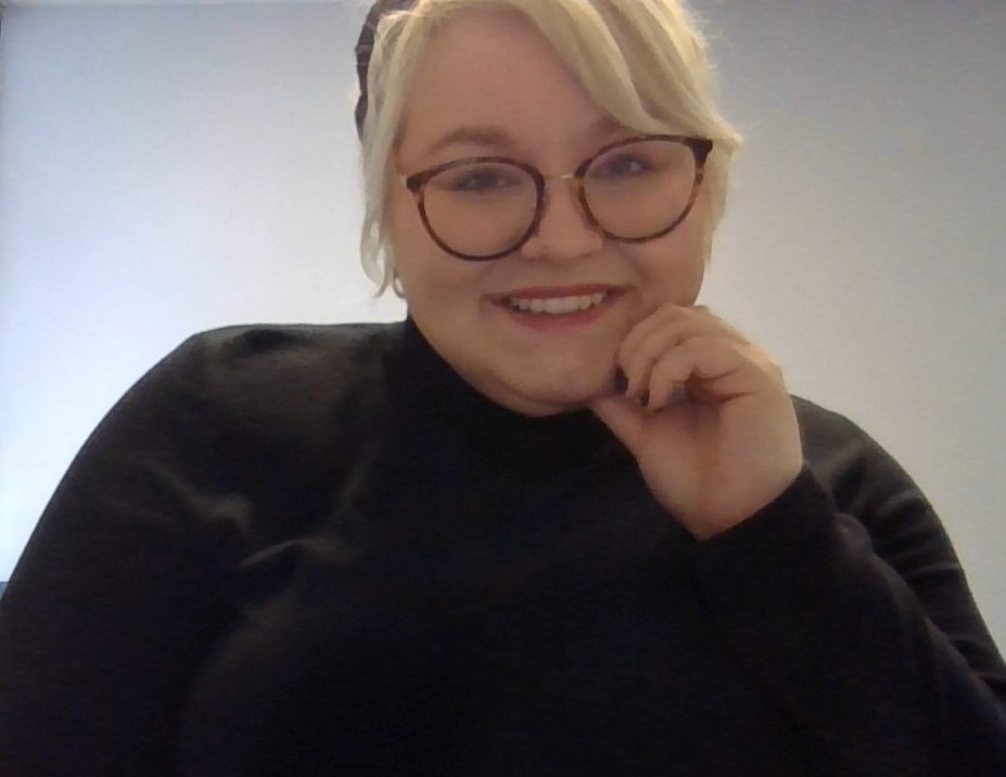 Photo of Jordan Hayes, a white young woman with dark framed glasses, blonde hair in an updo with bangs, red lipstick, and black long-sleeve shirt.