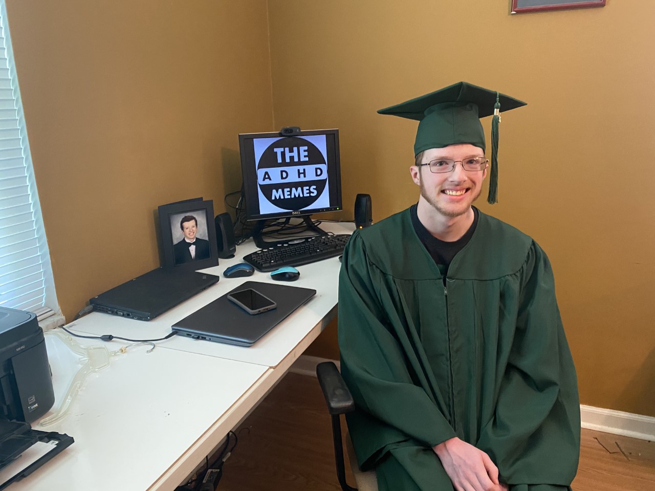 Photo of Zach Staubitz, a young white man with glasses. He is sitting at a desk wearing a green graduation cap and gown.